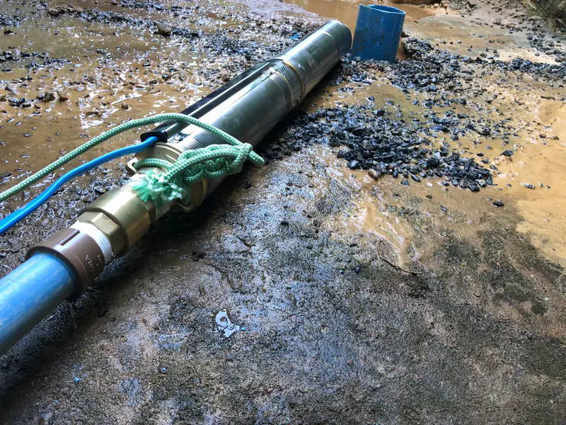 Bomba submersa metálica ligada a um tubo azul, com cabos e corda sobre terreno com brita.